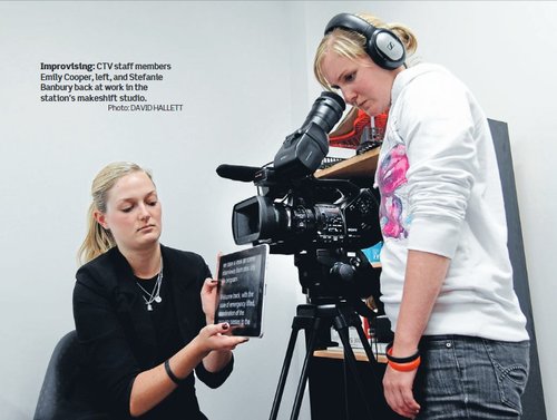 A photo of two staff members of Canterbury Television. One has large headphones on and looks down through the lens of a video camera. The other staff member sits on a chair, holding a screen with text just under the camera, for the TV presenters to read from.