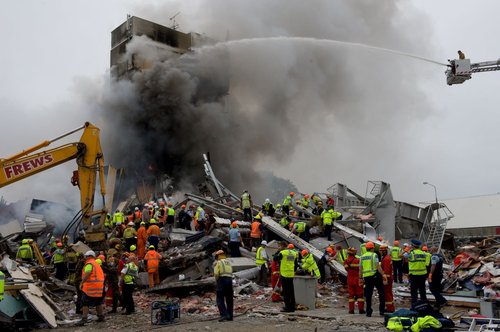 A member of the New Zealand Fire Service in a cherry picker spraying water at the fire burning in the collapsed Canterbury Television Building. Smoke is billowing out of the intact section of the building. Below, emergency personnel are searching the rubble for trapped people. A piece of corrugated plastic is being used to slide pieces of debris off the site.