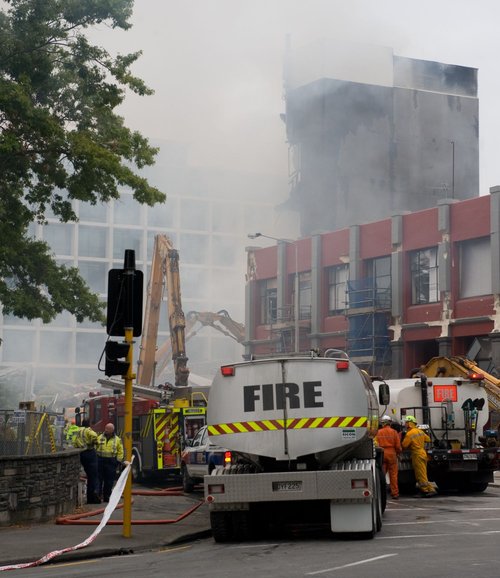 Smoke issuing from the collapsed Canterbury Television building on Madras Street. Fire Service personnel have gathered around the site to control the fire. In the background, excavators can be seen digging through the rubble.