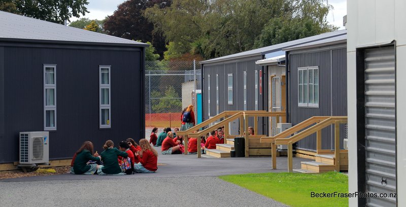 A photograph of Avonside Girls High School at morning tea time. Groups of girls sit in circles on the asphalt, in betweenblack-clad portacom temporary buildings.