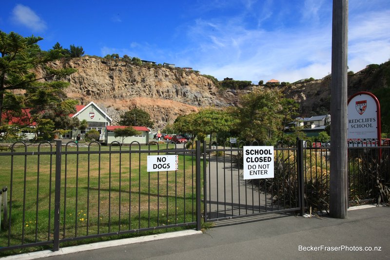 A photograph of Redcliffs School from outside the gate. On the fence are two signs - on that says "No dogs," and the other that reads, "School closed, do not enter." Behind them, some school buildings are visible, as well as the collapsed cliffs behind them.