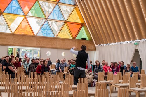 A photograph of people gathered in the Christchurch Transitional Cathedral, otherwise known as the Cardboard Cathedral. Johnny McFarlane of Beca is talking about project management of transitional architecture. The talk was part of a tour of the Cathedral, during FESTA 2013.