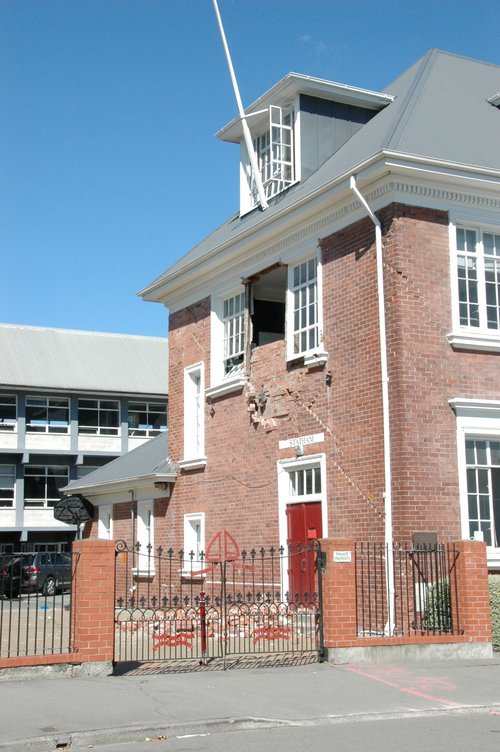 A photograph of the old red-brick Statham block at the Cathedral Grammar School. Between two windows, the wall from the upstairs floor has collapsed. Bricks are just visible on the ground behind the closed gate. Other cracks are visible on the building.