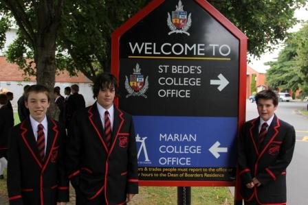 Three St Bede&#x27;s boys in uniform standing beside the welcome sign at their school. The sign is split top-and-bottom into two sections. The top section is black, reads "St Bede&#x27;s College Office" with their school logo and an arrow pointing to the right. The bottom half is blue, and reads "Marian College Office" with their school logo and an arrow pointing to the left.